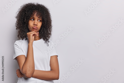 Unhappy frustrated young African woman with dark curly hair keeps hand under chin looks sadly aside ressed in casual t shirt has bad mood dressed in casual t shirt isolated over white background