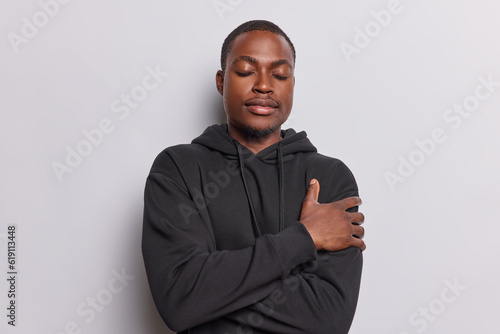 Horizontal shot of dark skinned man dressed in casual black sweatshirt embraces himself his eyes closed in state of calm and contentment finds comfort in warmth of his own embrace isolated on white
