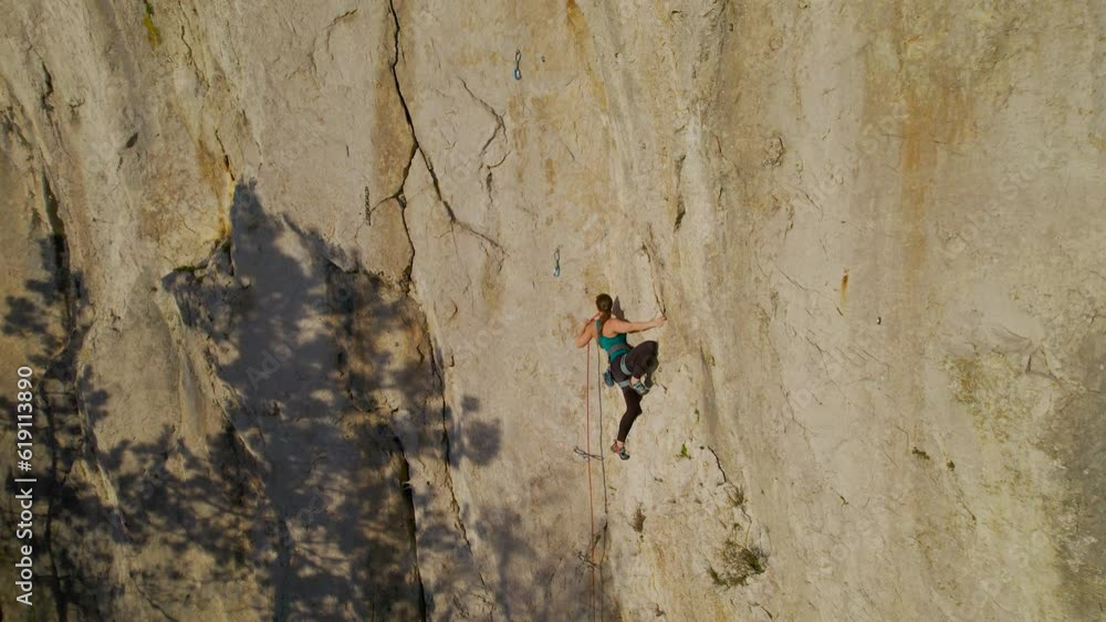 AERIAL: Female climber in the middle of a climb on a difficult climbing route. She is clipping in belay rope while lead climbing in limestone wall at incredible climbing playground in Karst landscape. AERIAL: Female climber in the middle of a climb on a difficult climbing route. She is clipping in belay rope while lead climbing in limestone wall at incredible climbing playground in Karst landscape.