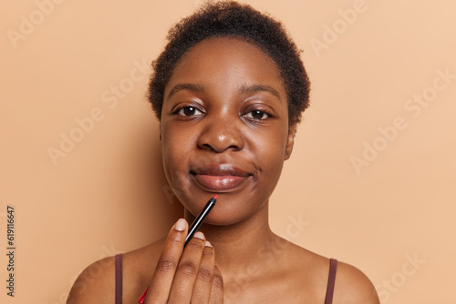 Horizontal shot of pretty curly haired young woman paints lips holds lip pencil does makeup stands bare shouldered looks directly at camera isolated over brown background. Beauty procedures.