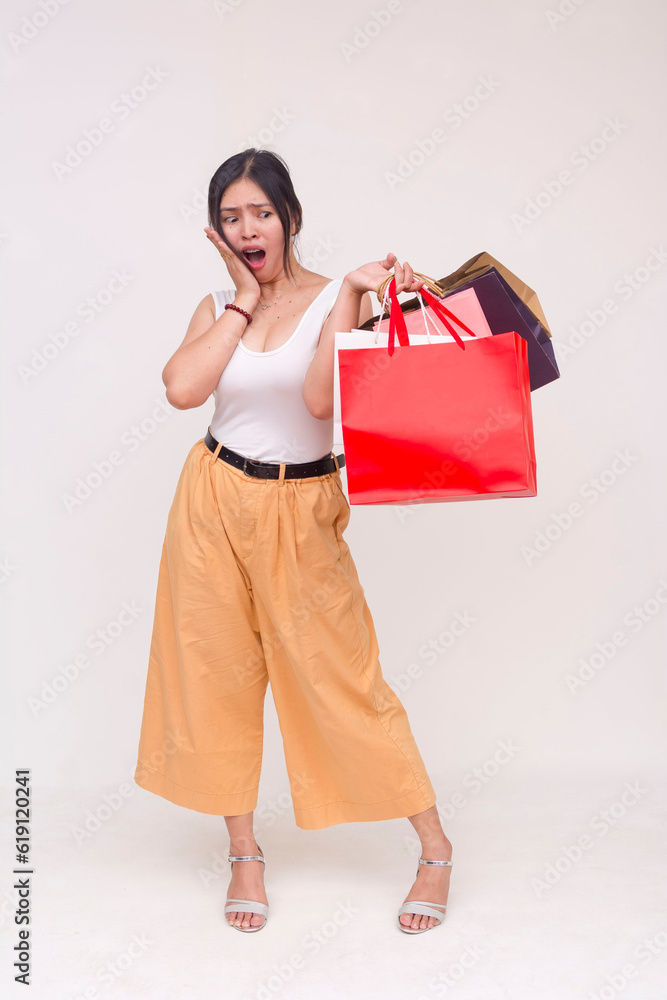 A shocked young Asian woman surprised at the multiple shopping bags she's holding in one hand. Isolated on a white background.