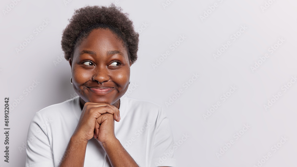 Portrait of dreamy dark skinned woman keeps hands under chin concentrated aside thinks about something pleasant dressed in casual t shirt poses against white background with copy space for your promo Portrait of dreamy dark skinned woman keeps hands under chin concentrated aside thinks about something pleasant dressed in casual t shirt poses against white background with copy space for your promo