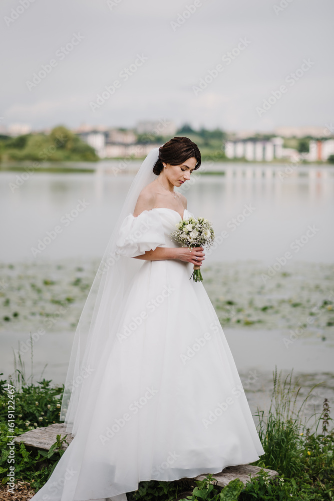 Young beautiful bride in a dress on shore of a lake outdoors. Happy wedding day of marriage. Getting married outdoors. Portrait of bride holding a bouquet white flowers roses and gypsophila in nature. Young beautiful bride in a dress on shore of a lake outdoors. Happy wedding day of marriage. Getting married outdoors. Portrait of bride holding a bouquet white flowers roses and gypsophila in nature.