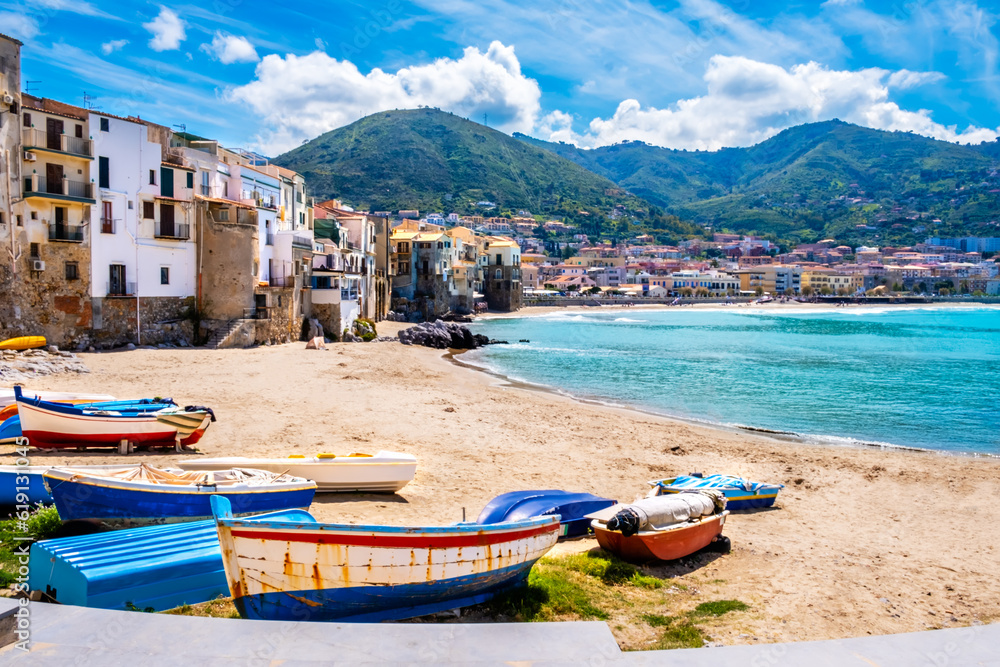 Fishing boats on beach of Cefalu, medieval town on Sicily island, Italy. Seashore village with historic buildings, clear turquoise sea water and mountains. Popular tourist attraction near Palermo