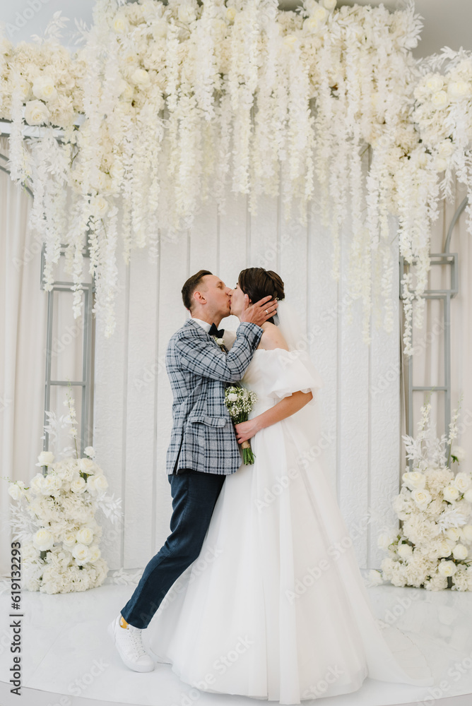 The bride and groom kissing. Newlyweds with a bouquet, standing on wedding ceremony under arch decorated white flowers. Full length of attractive woman wearing dress and man while ceremony. Marriage. The bride and groom kissing. Newlyweds with a bouquet, standing on wedding ceremony under arch decorated white flowers. Full length of attractive woman wearing dress and man while ceremony. Marriage.