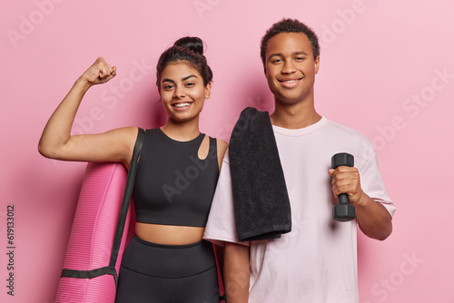 Horizontal shot of sporty couple trains with dumbbells show biceps carry karemat stand closely to each other dressed in sporswear isolated over pink background. People and healthy lifestyle concept