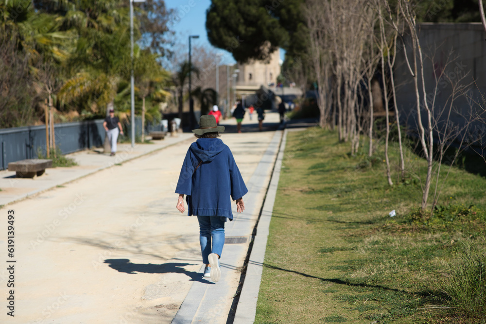 Senior woman is walking along a path with trees on either side. The woman is dressed in blue and wearing a hat.