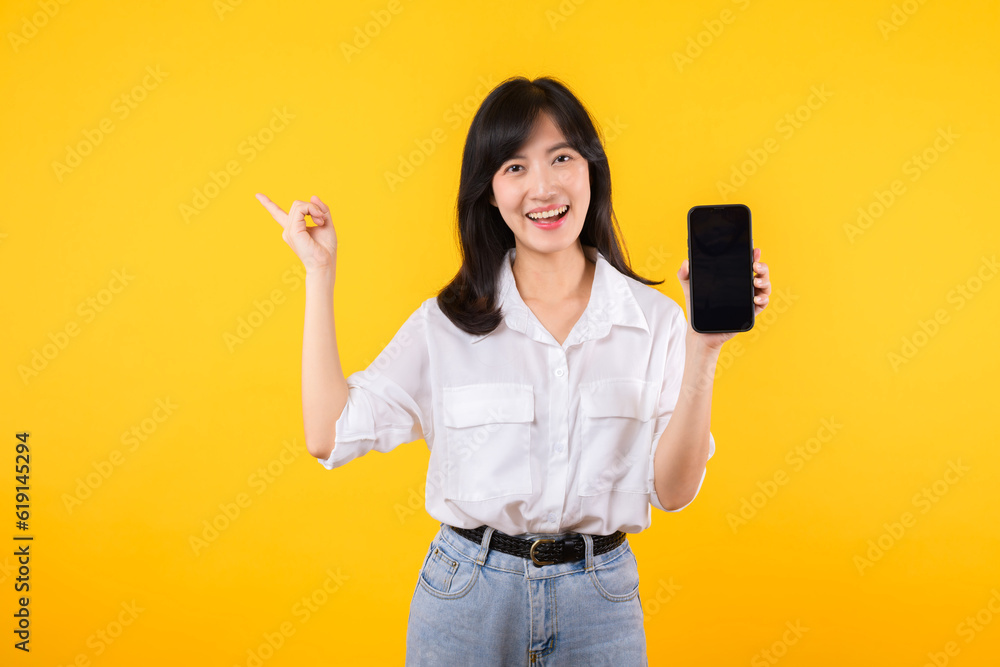 Portrait beautiful asian businesswoman happy woman wearing white shirt and denim plants showing blank smartphone screen and winner success body gesture feeling with fist up against yellow background. Portrait beautiful asian businesswoman happy woman wearing white shirt and denim plants showing blank smartphone screen and winner success body gesture feeling with fist up against yellow background.