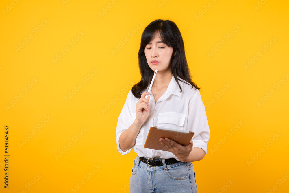 Doubtful Asian young adult business woman working on digital tablet and looking away thinking of solution isolated on yellow background, considering offer, making decision or feeling doubt concept. Doubtful Asian young adult business woman working on digital tablet and looking away thinking of solution isolated on yellow background, considering offer, making decision or feeling doubt concept.