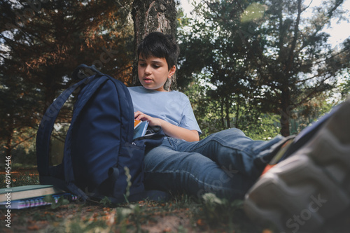 Smart teenage schoolboy sitting under a tree, opening his backpack, taking out textbook for doing homework outdoors