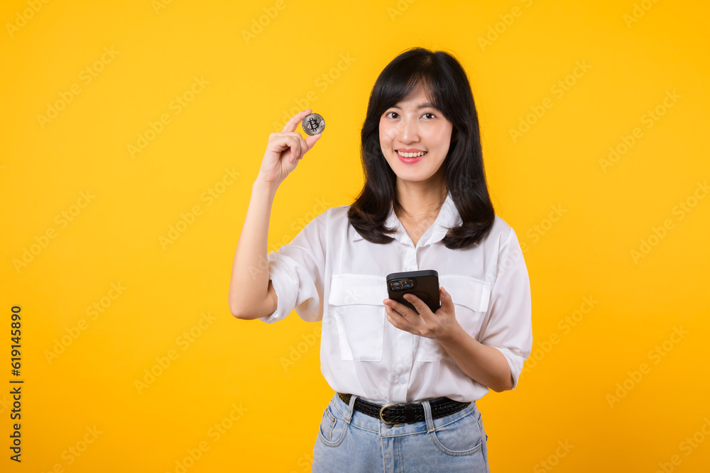 Stunning portrait of an Asian young woman holding digital currency and smartphone on yellow studio background. Explore the concept of crypto digital currency. Stunning portrait of an Asian young woman holding digital currency and smartphone on yellow studio background. Explore the concept of crypto digital currency.