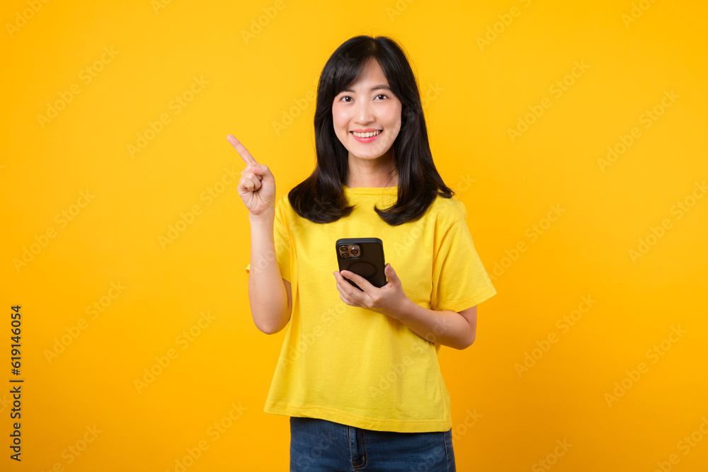Capture young Asian woman wearing a yellow t-shirt and denim jeans points with finger hand gesture to free copy space while using smartphone. Perfect for illustrating the concept of app functionality. Capture young Asian woman wearing a yellow t-shirt and denim jeans points with finger hand gesture to free copy space while using smartphone. Perfect for illustrating the concept of app functionality.