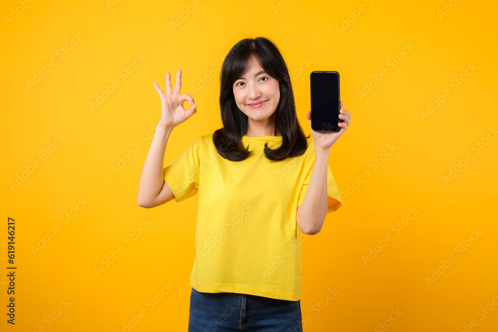 positive portrait young Asian woman wearing yellow t-shirt and denim jeans showing happy smile while showing smartphone screen and making the okay hand sign gesture. recommended applications concept. positive portrait young Asian woman wearing yellow t-shirt and denim jeans showing happy smile while showing smartphone screen and making the okay hand sign gesture. recommended applications concept.