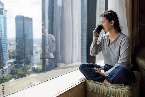 Young woman holding coffee cup, wearing pajama and looking at cityscape through the window in luxury penthouse apartment or hotel room