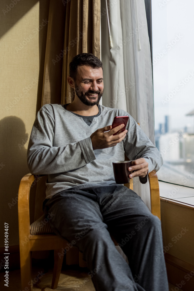 Young man holding coffee cup, wearing pajama and laughing looking at the smartphone. Man sitting by the window in luxury penthouse apartment or hotel room