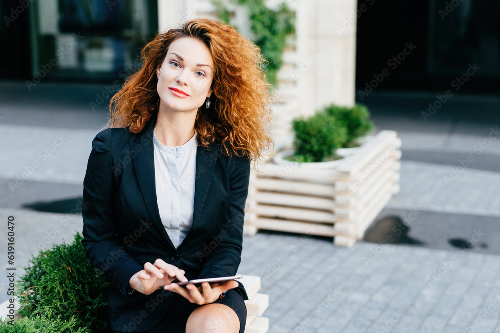 Pretty businesswoman with curly hair, red lips, formal attire, sitting at outdoor cafe, using Wi-Fi, socializing with friends.