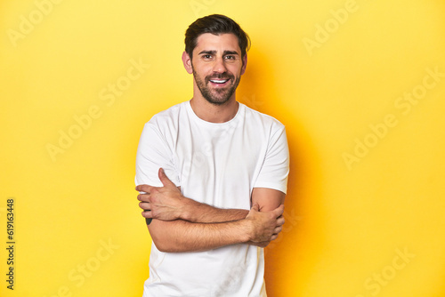 Caucasian man in white t-shirt on yellow studio background who feels confident, crossing arms with determination.