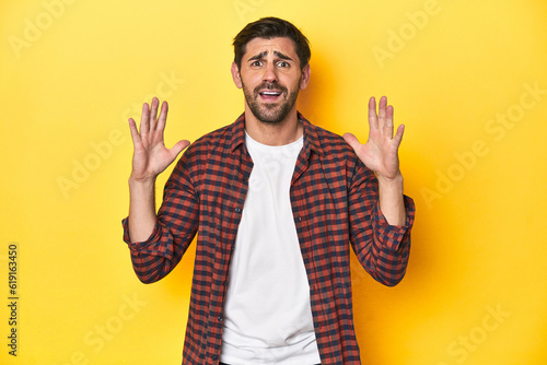 Caucasian man in red checkered shirt, yellow backdrop screaming to the sky, looking up, frustrated.