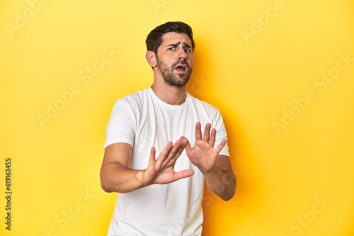 Caucasian man in white t-shirt on yellow studio background rejecting someone showing a gesture of disgust.