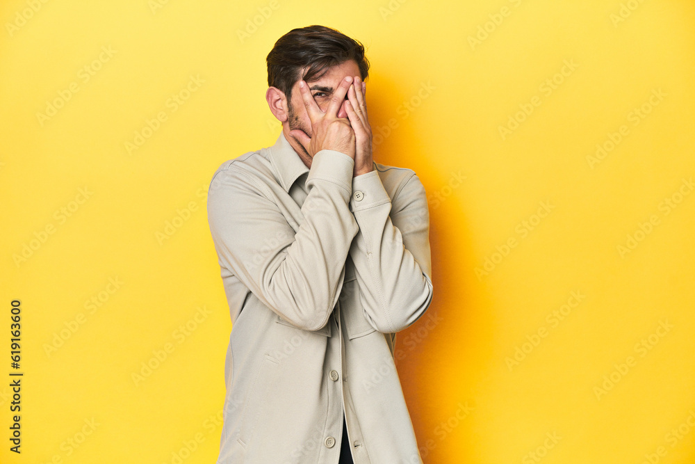 Caucasian man in grey shirt, studio shot on yellow blink through fingers frightened and nervous. Caucasian man in grey shirt, studio shot on yellow blink through fingers frightened and nervous.
