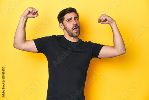 Caucasian man in black t-shirt, yellow studio backdrop showing strength gesture with arms, symbol of feminine power