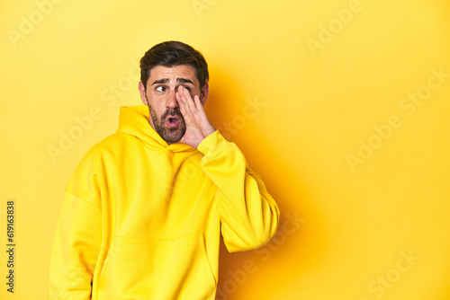 Man in yellow hoodie, monochrome studio backdrop is saying a secret hot braking news and looking aside © Asier