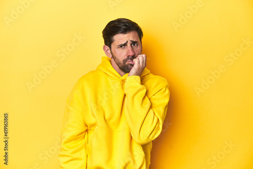 Man in yellow hoodie, monochrome studio backdrop biting fingernails, nervous and very anxious.