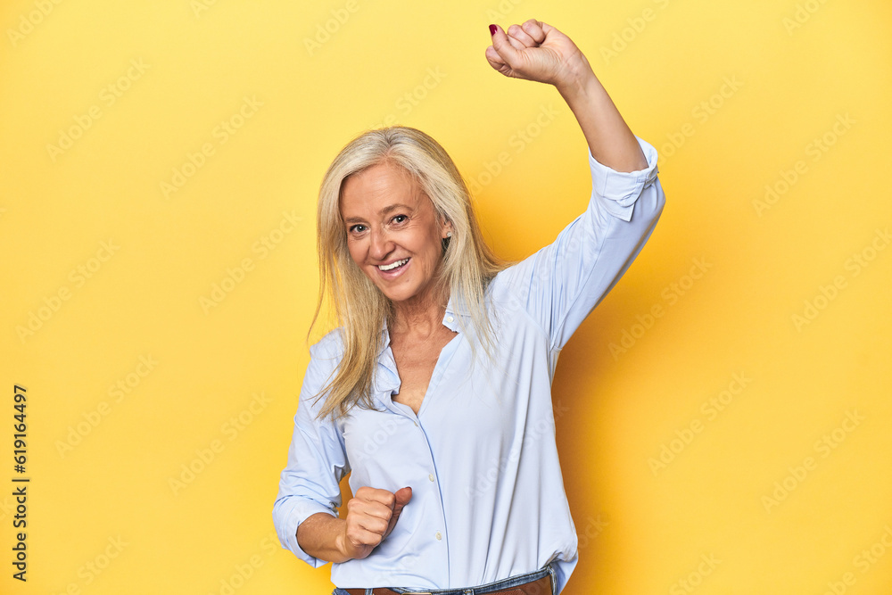 Middle-aged Caucasian woman in blue shirt, yellow studio celebrating a special day, jumps and raise arms with energy. Middle-aged Caucasian woman in blue shirt, yellow studio celebrating a special day, jumps and raise arms with energy.