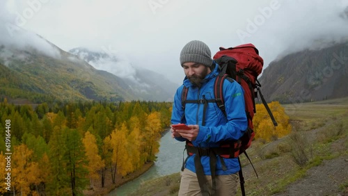 Caucasian backpacker with rucksack use smartphone online, hold device, touch, scroll and tap. Young male person in equipment at extreme vacation walk and use cellphone. Beautiful autumn natural area