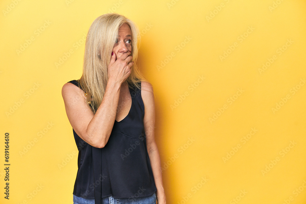 Elegant middle-aged woman in black top, yellow studio thoughtful looking to a copy space covering mouth with hand. Elegant middle-aged woman in black top, yellow studio thoughtful looking to a copy space covering mouth with hand.