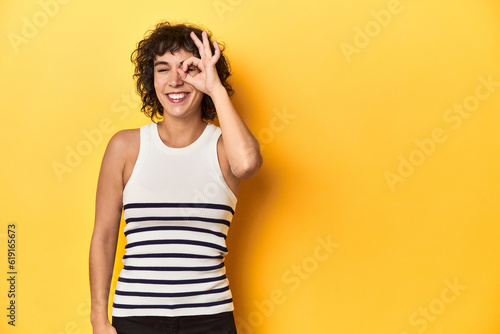 Caucasian curly-haired woman in white tank-top excited keeping ok gesture on eye.