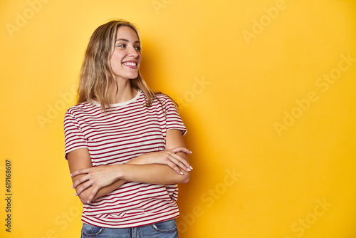 Young blonde Caucasian woman in a red striped t-shirt on a yellow background, smiling confident with crossed arms.
