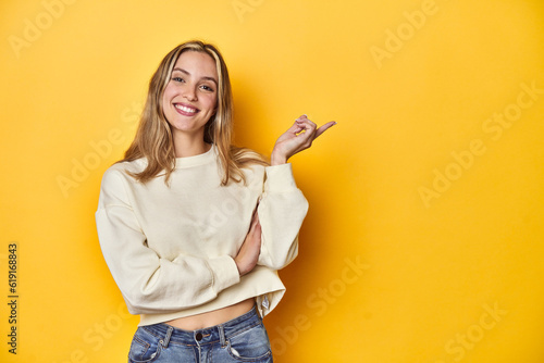 Young blonde Caucasian woman in a white sweatshirt on a yellow studio background, smiling cheerfully pointing with forefinger away.