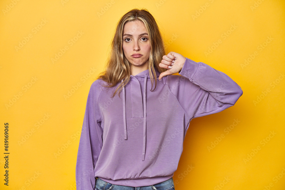 Young blonde Caucasian woman in a violet sweatshirt on a yellow background, showing a dislike gesture, thumbs down. Disagreement concept. Young blonde Caucasian woman in a violet sweatshirt on a yellow background, showing a dislike gesture, thumbs down. Disagreement concept.