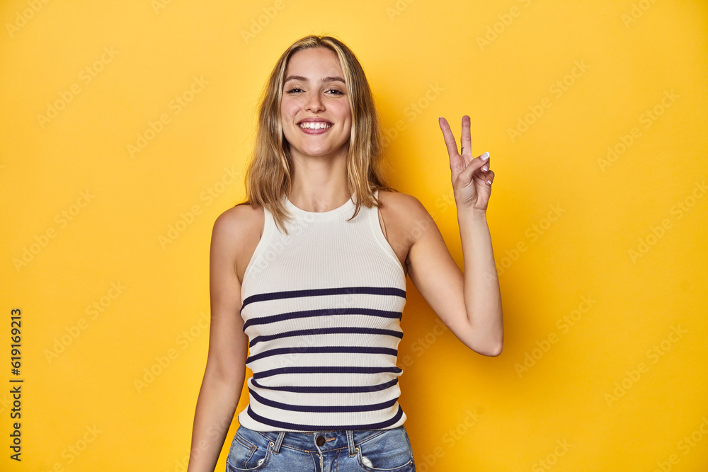 Young blonde Caucasian woman in a white tank top on a yellow studio background, joyful and carefree showing a peace symbol with fingers. Young blonde Caucasian woman in a white tank top on a yellow studio background, joyful and carefree showing a peace symbol with fingers.