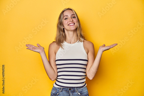 Young blonde Caucasian woman in a white tank top on a yellow studio background, makes scale with arms, feels happy and confident.