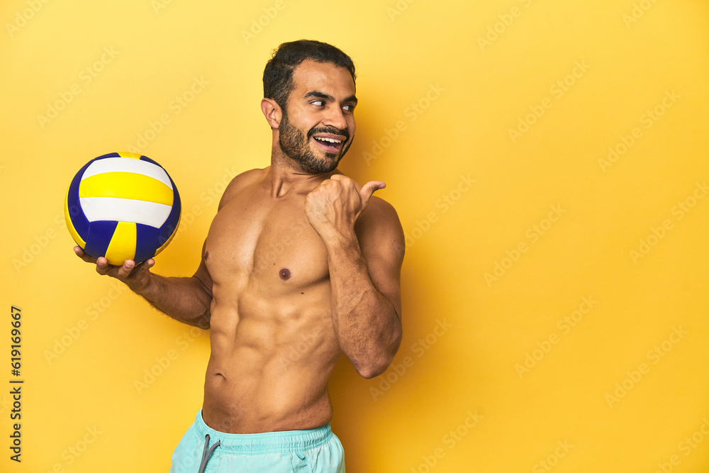 Young athletic Latino man, shirtless in swimwear, holding a volleyball points with thumb finger away, laughing and carefree.