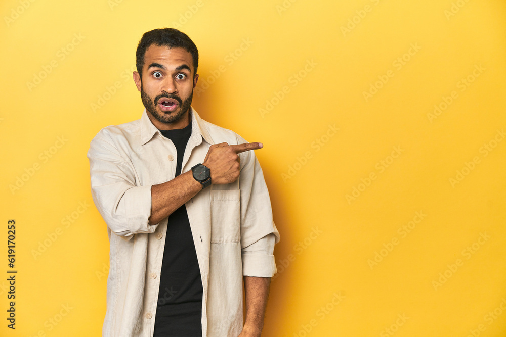 Casual young Latino man against a vibrant yellow studio background, pointing to the side Casual young Latino man against a vibrant yellow studio background, pointing to the side