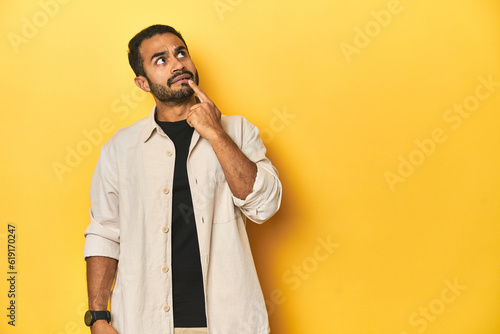 Casual young Latino man against a vibrant yellow studio background, looking sideways with doubtful and skeptical expression.