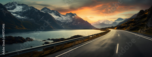 A scenic road winds alongside a serene lake, as vibrant clouds dance in the sunset-lit sky. Majestic mountains complete the breathtaking panorama, creating a picturesque and awe-inspiring view.