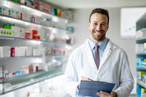 Portrait of caucasian male young pharmacist druggist in white medical coat holding clipboard with side effects, active substance, prescriptions standing at pharmacy drugstore photo