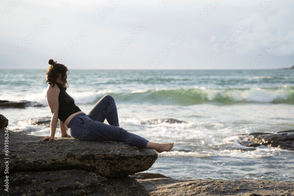 young girl sitting down comfortly on the rocks in the beach at sunset. Golden hour