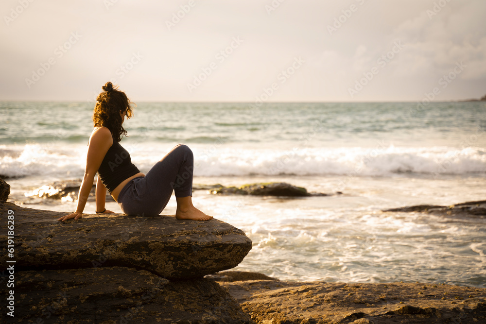young girl sitting down comfortly on the rocks in the beach at sunset. Golden hour