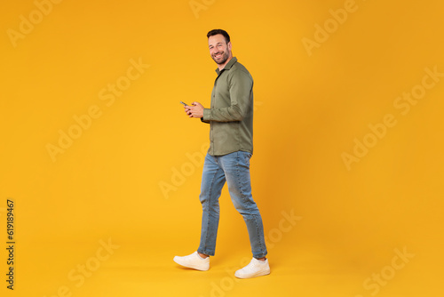 Full length shot of happy man walking with cellphone in hands and smiling at camera, messaging or shopping online on yellow background, side view, free space