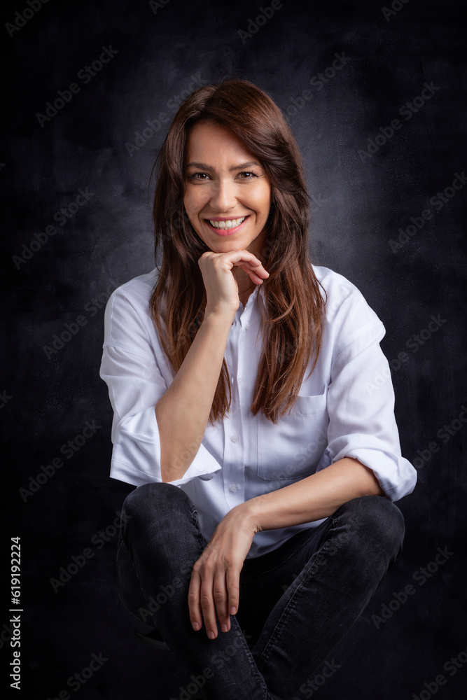 Studio portrait of a brunette haired attractive woman wearing white shirt and black jeans while posing at isolated dark background Studio portrait of a brunette haired attractive woman wearing white shirt and black jeans while posing at isolated dark background