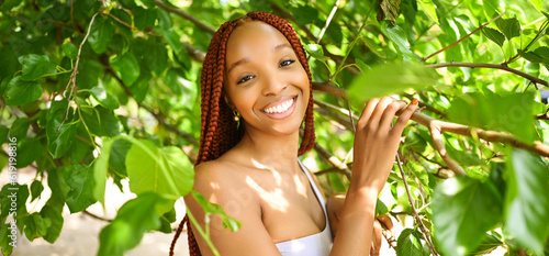 Close up portrait Beautiful young African American woman with red braids hair, perfect white teeth smiling outside, sunny summer day green foliage. Facial treatment. Cosmetology, skin care and spa