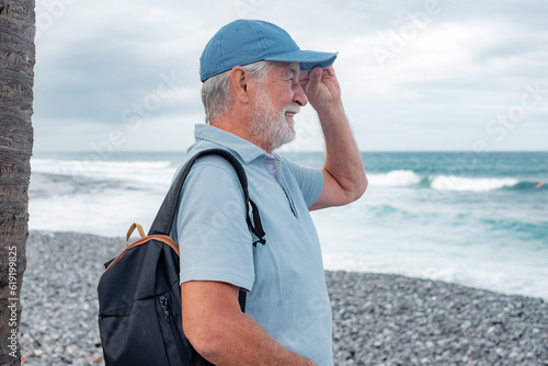 Side view of senior man in casual polo shirt standing at the beach looking at horizon over sea, pensioner male with cap enjoying free time and retirement #619199825