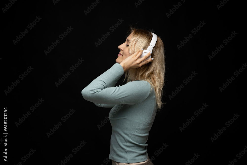 Profile portrait of charming blonde woman in massive white headphones listening to music on isolated black background. Profile portrait of charming blonde woman in massive white headphones listening to music on isolated black background.