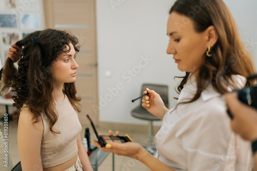 Medium shot of female makeup artist holding in hands eye shadow palette during applying eyeshadow in beauty salon to sensual young woman with curly hair. Concept of beauty and stylish makeup.