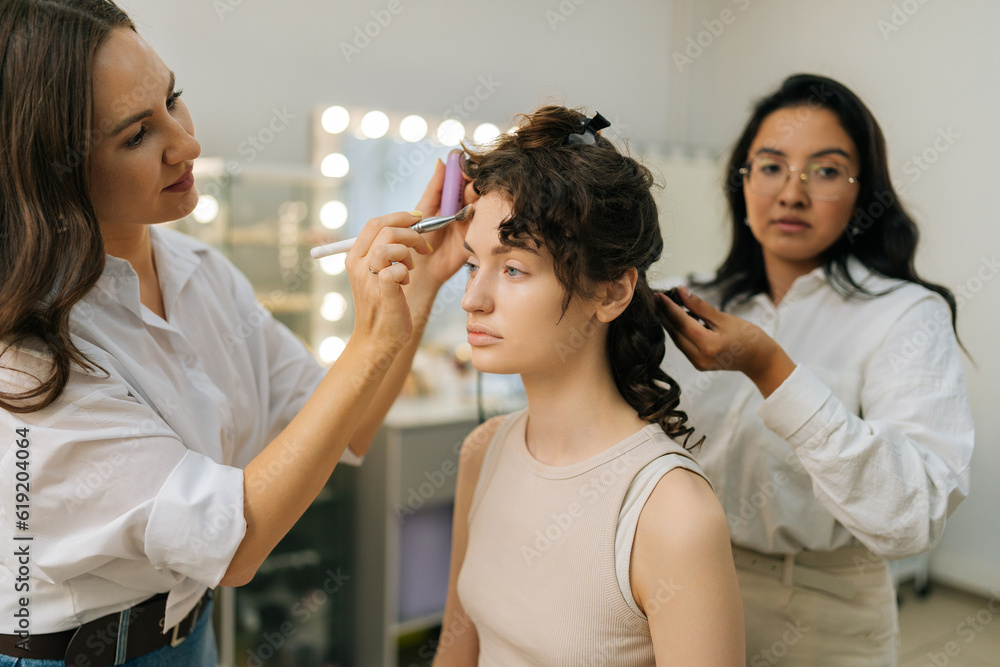 Portrait of pretty young woman being given applying highlighter for facial sculpting from makeup artist. Hairstylist doing stylish hairdo in beauty studio. Concept of make up and styling in 4 hands.